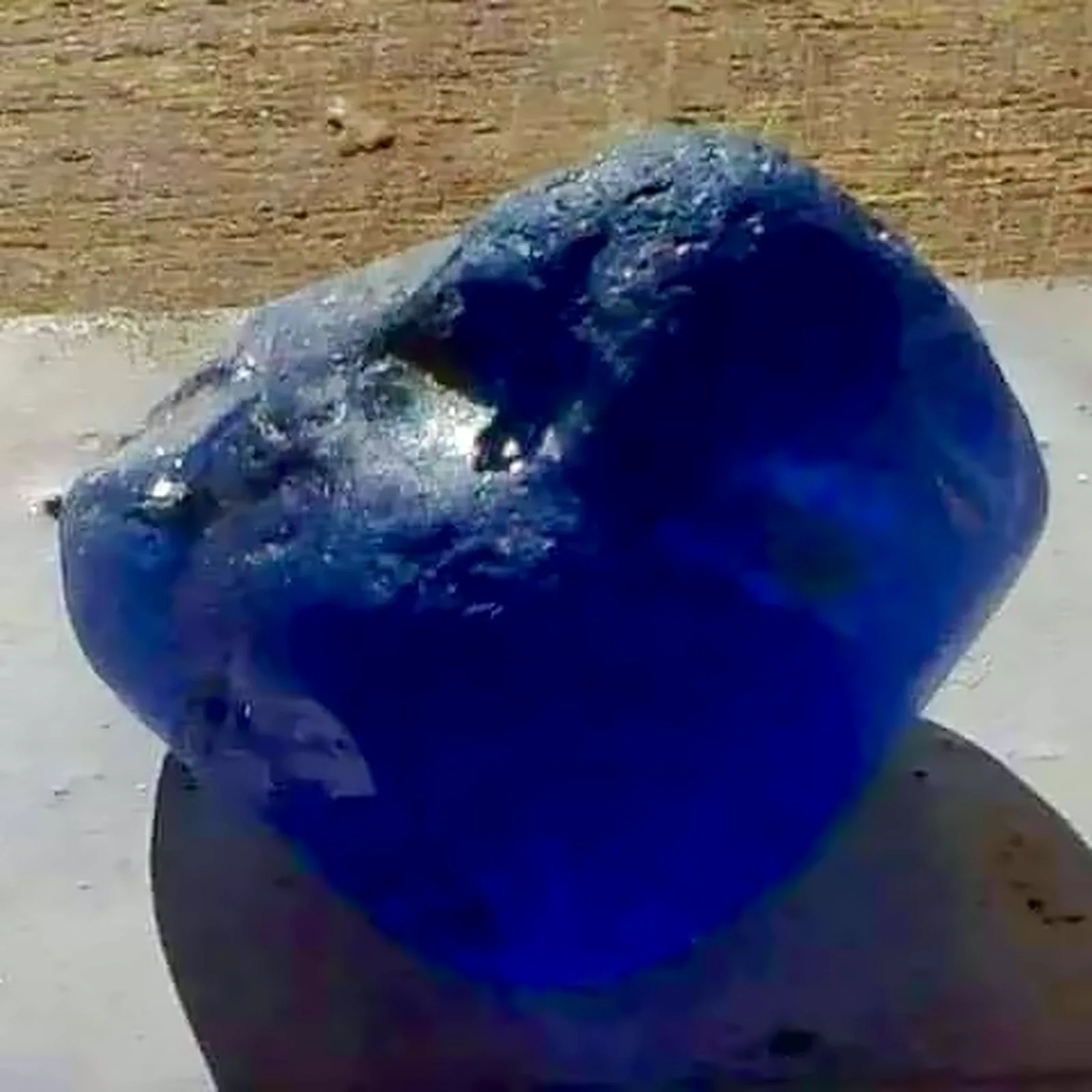 A single deep blue rough sapphire resting on a white surface in daylight.